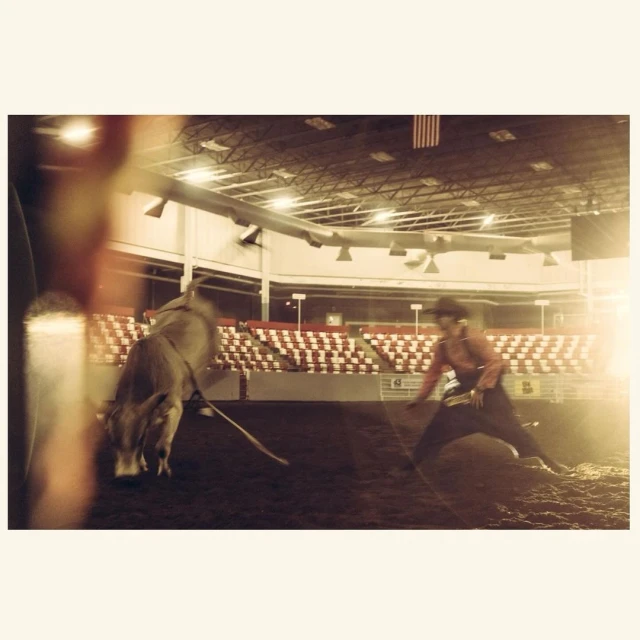 Rodeo action shot featuring a cowboy riding a bull in an indoor arena under bright lights, with an American flag in the background.