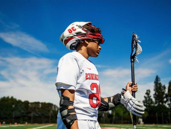 Young lacrosse player in helmet and pads holds his stick on a sports field under a blue sky with scattered white clouds.
