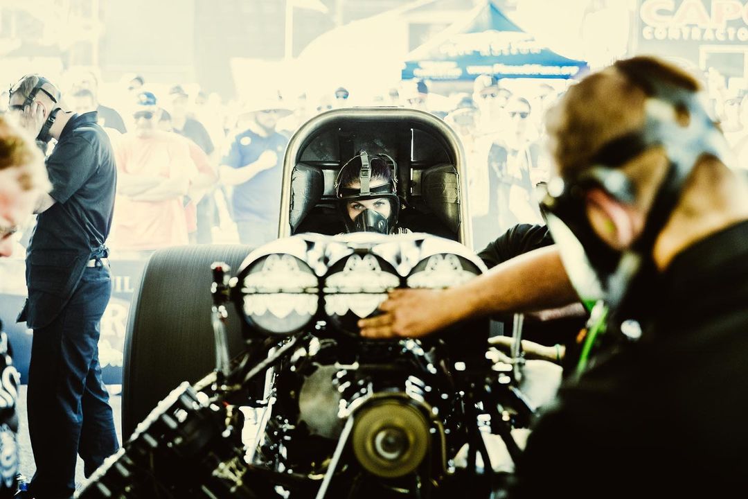 Drag racer wearing a helmet and mask in the cockpit of a race car at an outdoor event, prepared for a high-speed race.