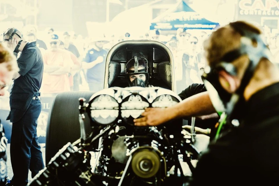 Drag racer wearing a helmet and mask in the cockpit of a race car at an outdoor event, prepared for a high-speed race.