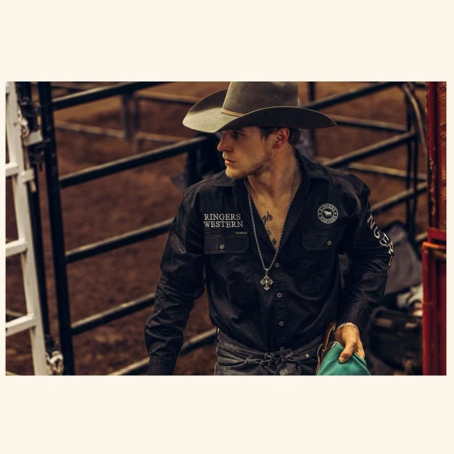 Cowboy in Ringers Western shirt, hat, and pendant poses in rodeo arena; casual, confident style showcases modern western fashion.