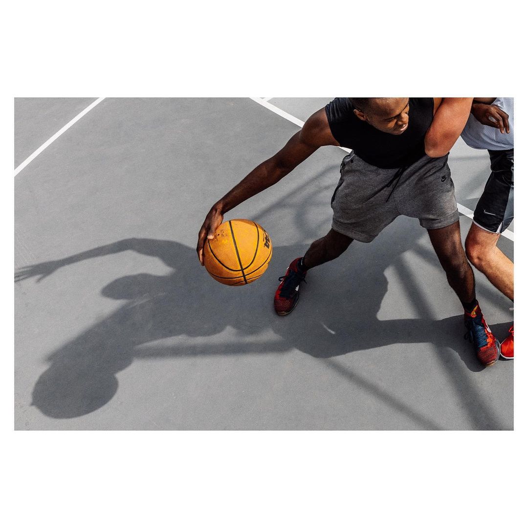 Two adult male basketball players competing on an outdoor court. One player is dribbling the ball, with his shadow cast on the grey surface.