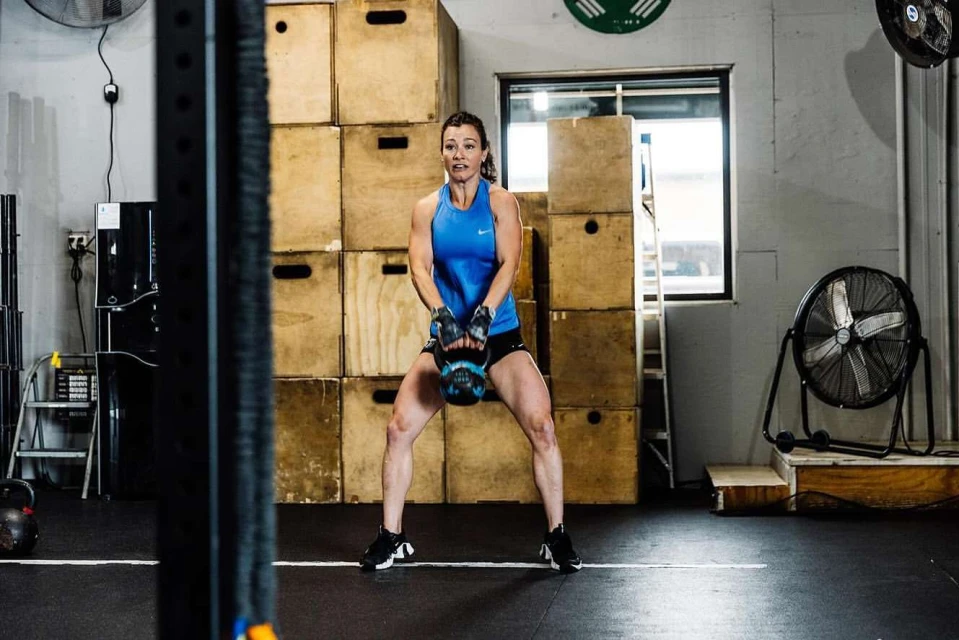 Athletic woman exercising with kettlebell in a fitness center. She wears sportswear, with weightlifting equipment and lockers in the background.