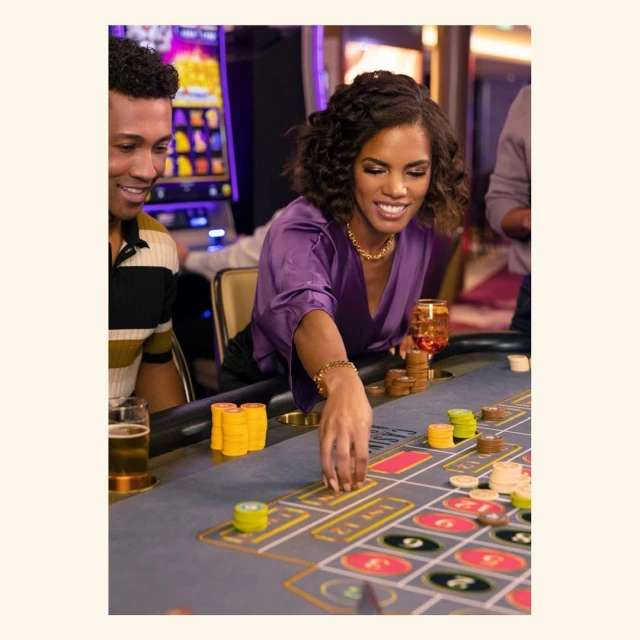 A woman in a purple blouse places chips on a roulette table in a casino, with a smiling man and slot machines visible in the background.