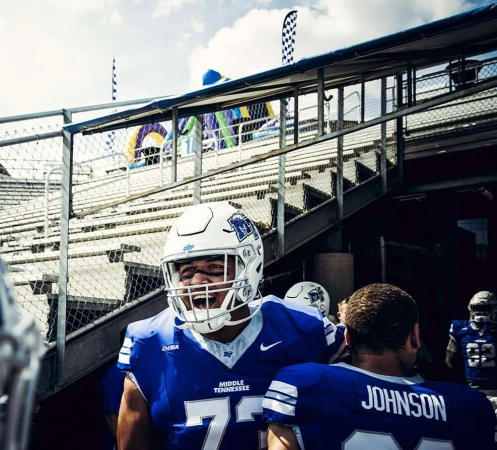 American football players in blue and white uniforms exiting the stadium. One player in the foreground is shouting with excitement.
