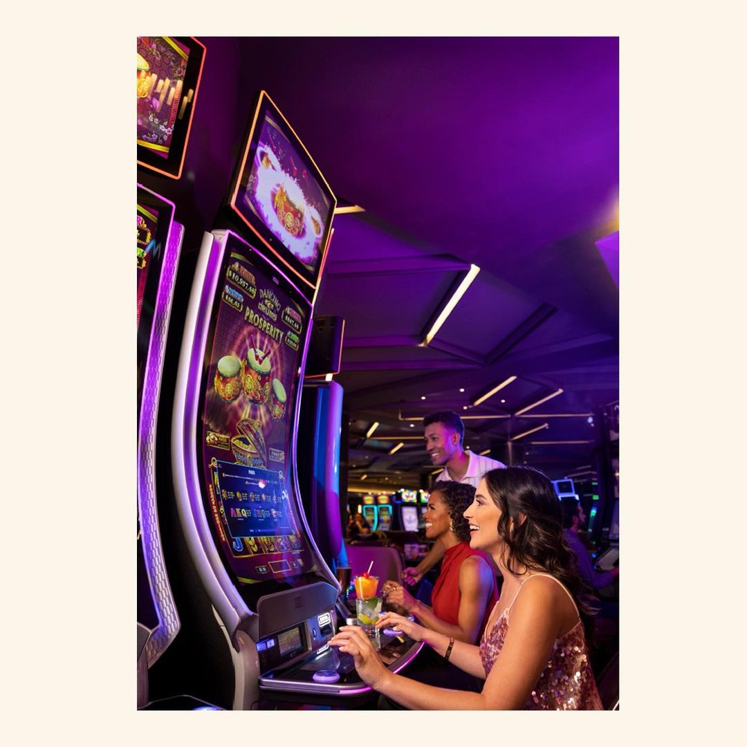 Slot machines glow with vibrant colors as three people play in a casino setting with purple ambient lighting, creating a lively atmosphere.
