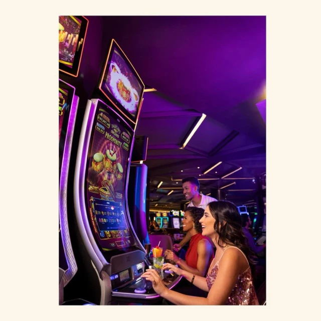 Slot machines glow with vibrant colors as three people play in a casino setting with purple ambient lighting, creating a lively atmosphere.