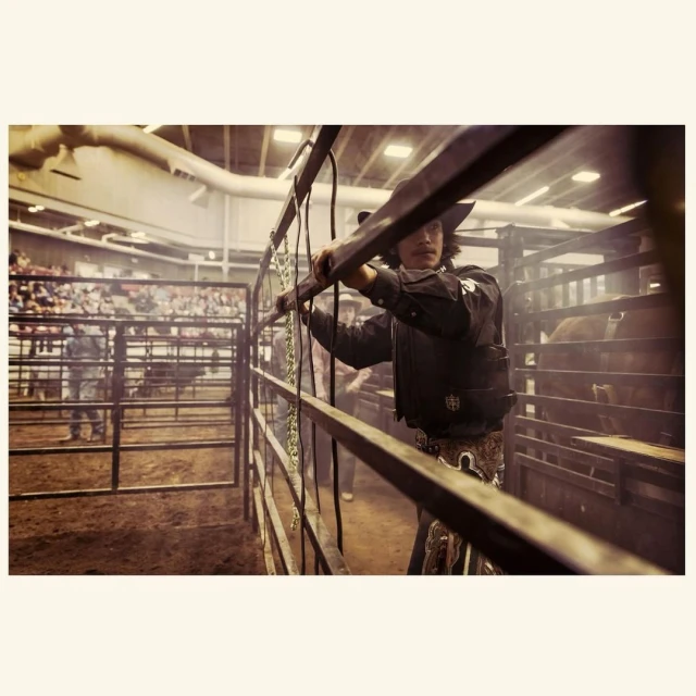 Cowboy stands inside the rodeo arena. Wearing a black shirt, vest, and cowboy hat, the man rests his arms on the railing.