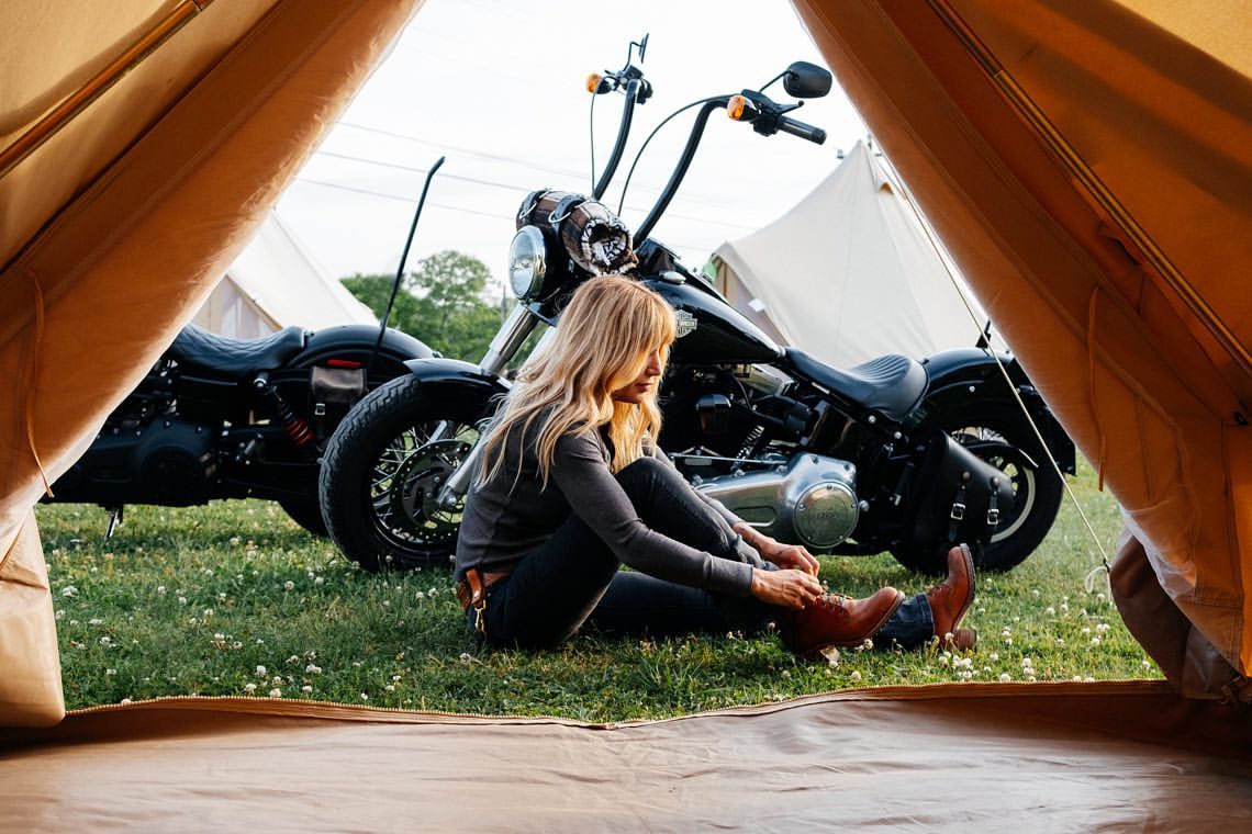A woman in jeans and a gray top sits on the grass tying her brown boots next to two black motorcycles near a tan canvas tent.