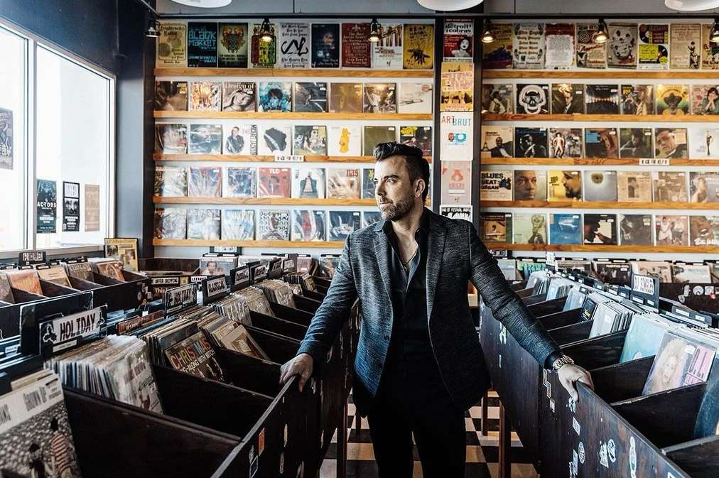 Man in grey jacket stands in a record store, surrounded by shelves and bins overflowing with vinyl records, browsing through the selection.