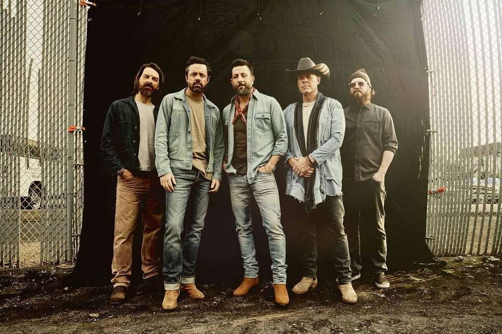 Group shot of five adult men in casual denim and western wear standing outdoors against a dark backdrop and chain-link fence.