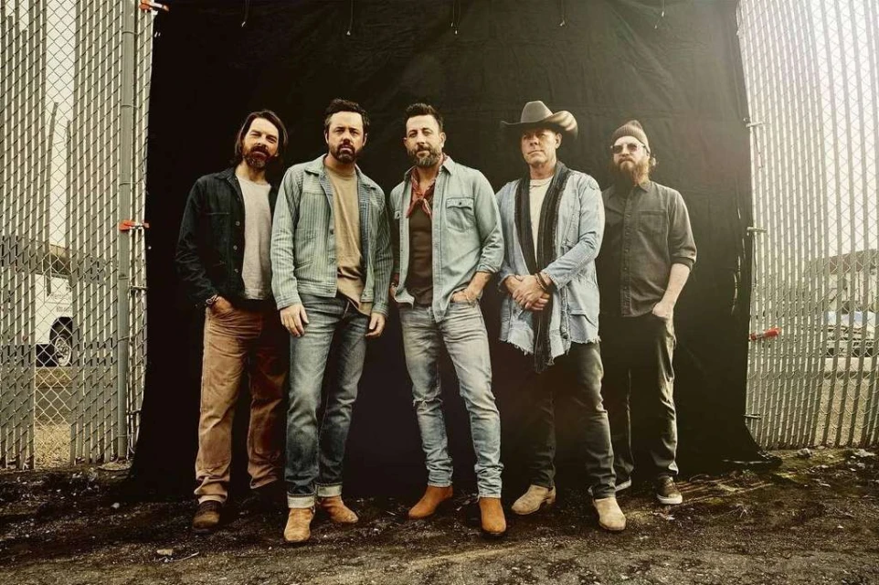 Group shot of five adult men in casual denim and western wear standing outdoors against a dark backdrop and chain-link fence.