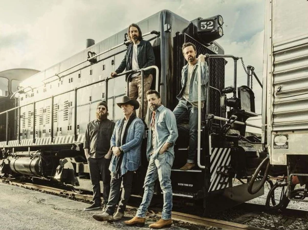 Five adult men stand in front of a vintage black train on train tracks in an outdoor setting, wearing casual clothing.