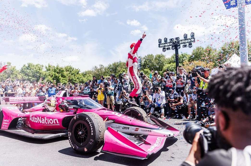 IndyCar race winner stands triumphantly atop his pink race car, surrounded by celebrating fans and photographers on the track.