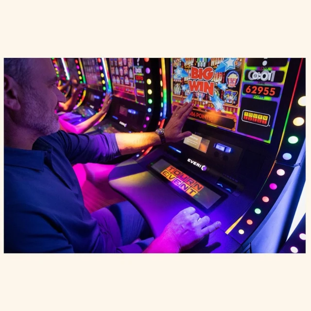 Man playing a slot machine with a vibrant screen displaying 'Big Win' and colorful lights in a casino, wearing blue shirt.