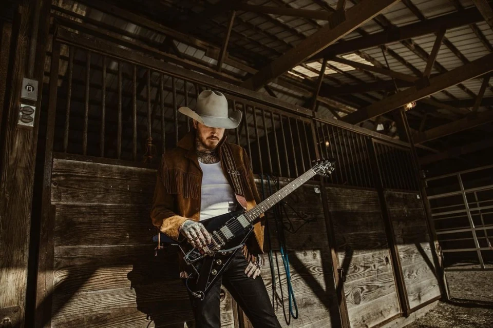 Man in cowboy hat and fringe jacket plays guitar inside a rustic barn. Dark and moody lighting adds to the visual atmosphere.