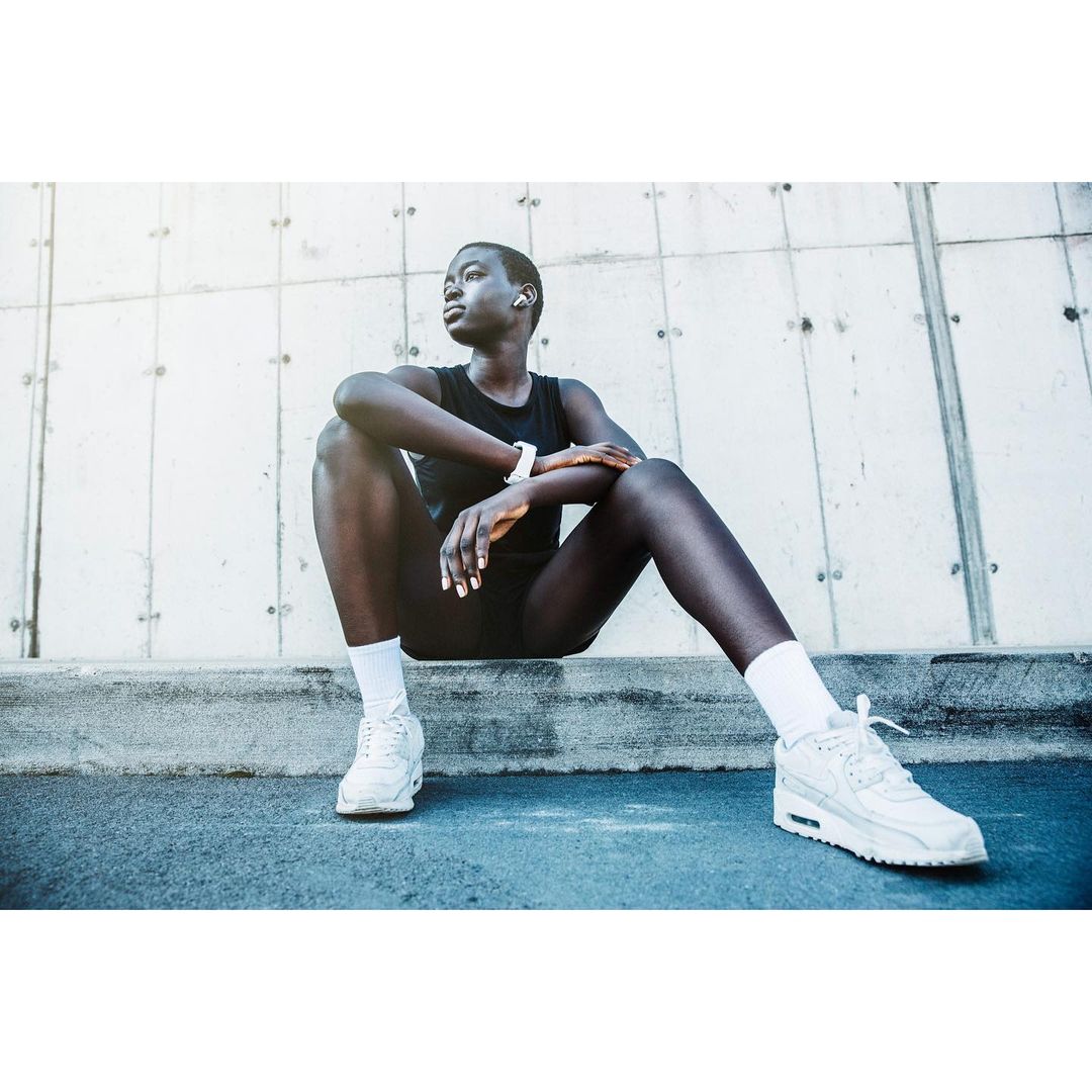 Athletic Black woman in sportswear sits outside on concrete, wearing sneakers, wireless earbuds and a smartwatch, looking off-camera.