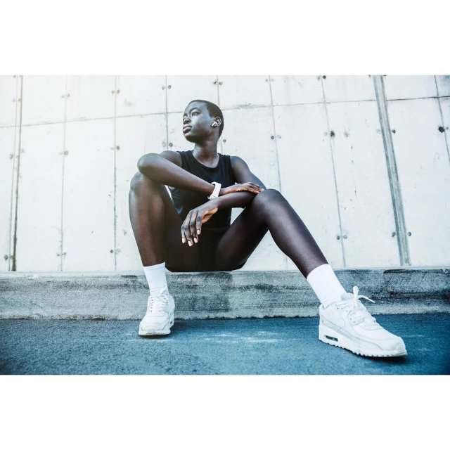 Athletic Black woman in sportswear sits outside on concrete, wearing sneakers, wireless earbuds and a smartwatch, looking off-camera.