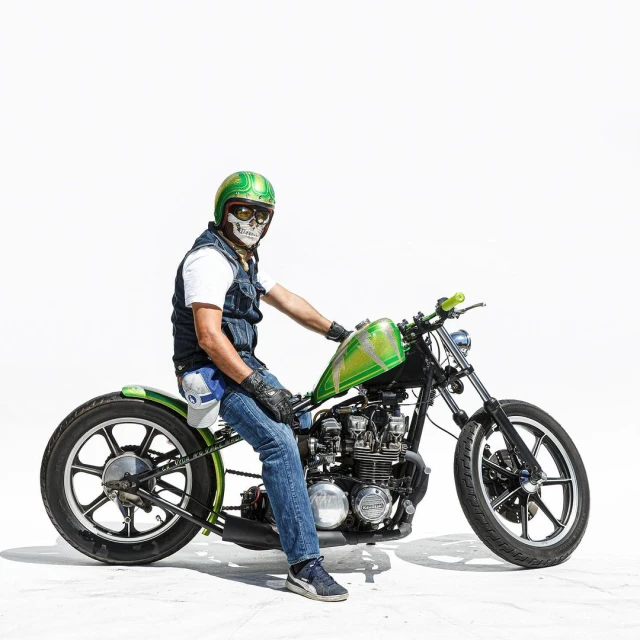 Adult male biker wearing a skull mask and helmet sits on a custom green and black motorcycle, shot in a bright photo studio setting.