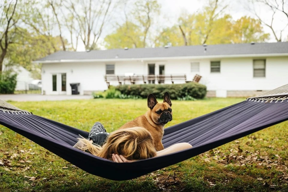Woman relaxes in a hammock with a French bulldog in a backyard with green grass and a white house during daytime under an overcast sky.