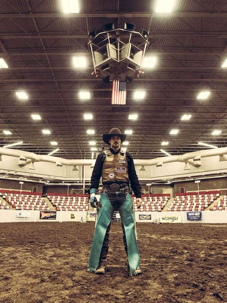 Cowboy stands in rodeo arena with hat, vest, and chaps. American flag hangs overhead in an indoor venue with stadium seating.