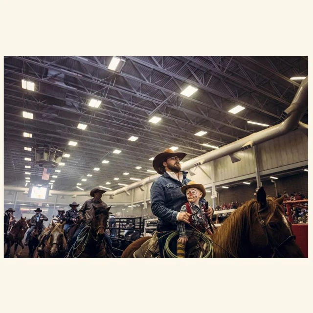 Group of cowboys and cowgirls on horseback indoors in a rodeo arena under bright lights, one cowboy holds a ventriloquist dummy.