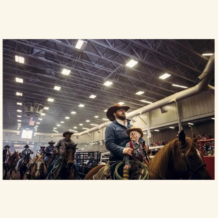 Group of cowboys and cowgirls on horseback indoors in a rodeo arena under bright lights, one cowboy holds a ventriloquist dummy.
