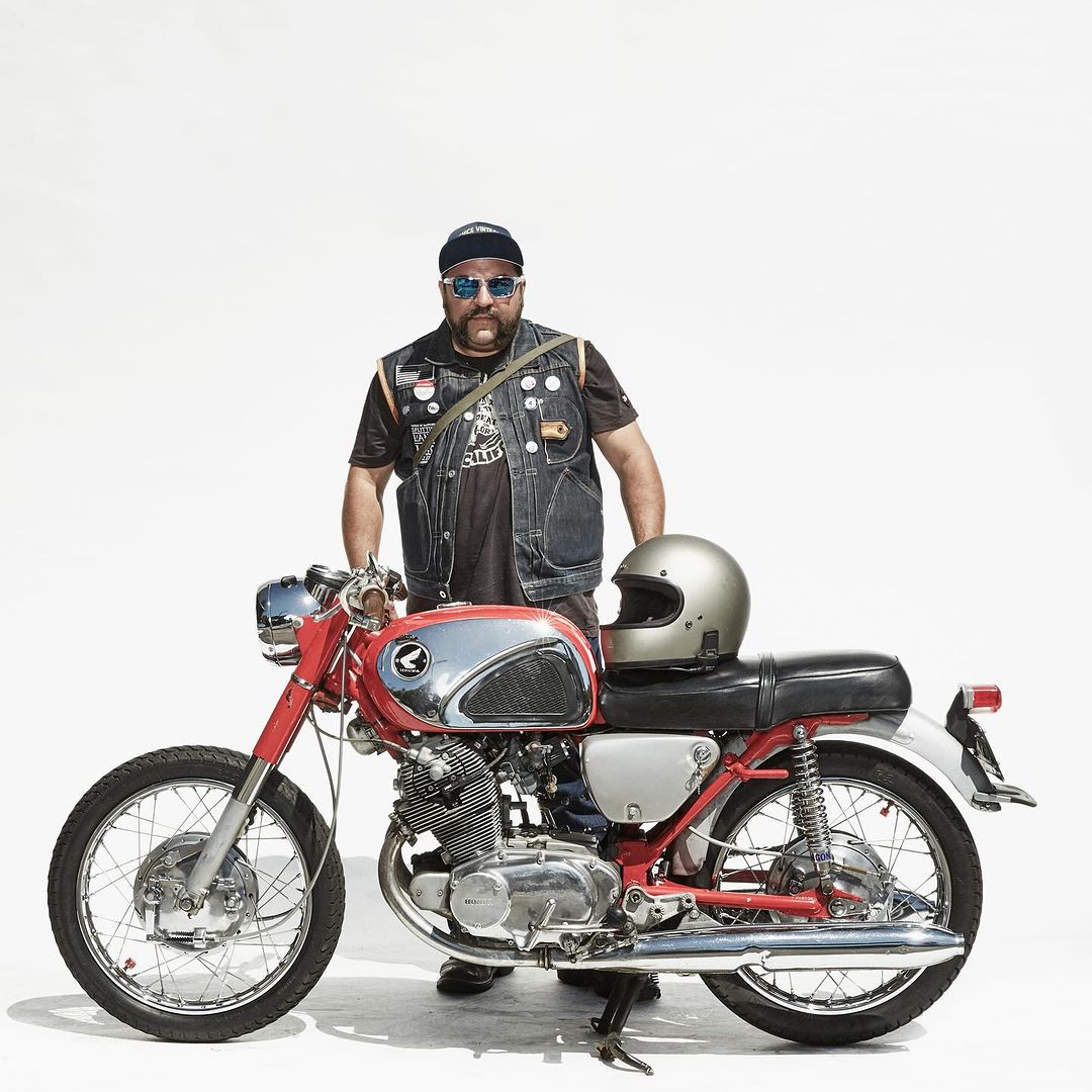 An adult male with a beard and sunglasses standing with a vintage motorcycle on a white background, wearing a denim vest and cap.