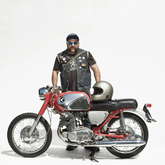 An adult male with a beard and sunglasses standing with a vintage motorcycle on a white background, wearing a denim vest and cap.