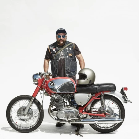 An adult male with a beard and sunglasses standing with a vintage motorcycle on a white background, wearing a denim vest and cap.