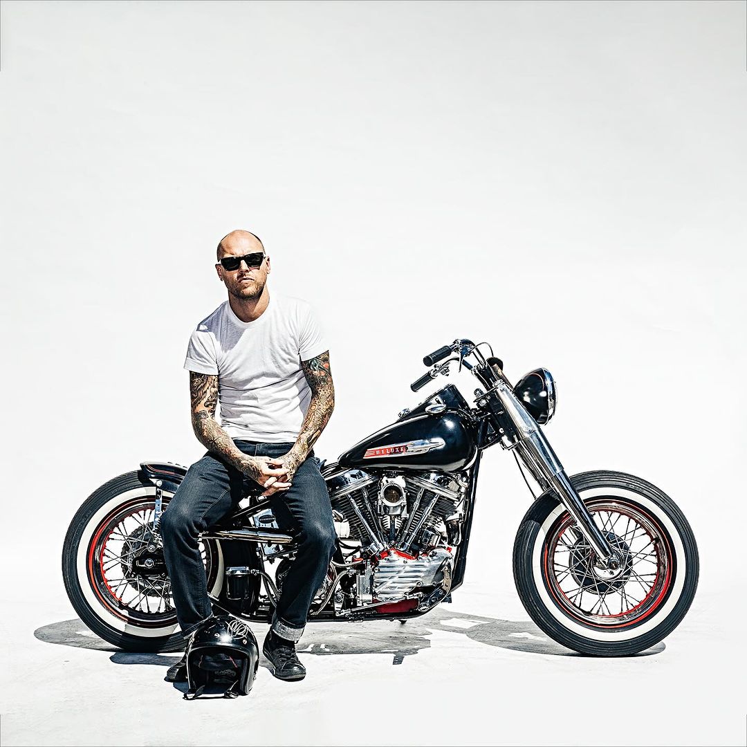 Bald adult male with tattoos sits on a vintage black motorcycle in a bright studio against a white background with a helmet on the ground.