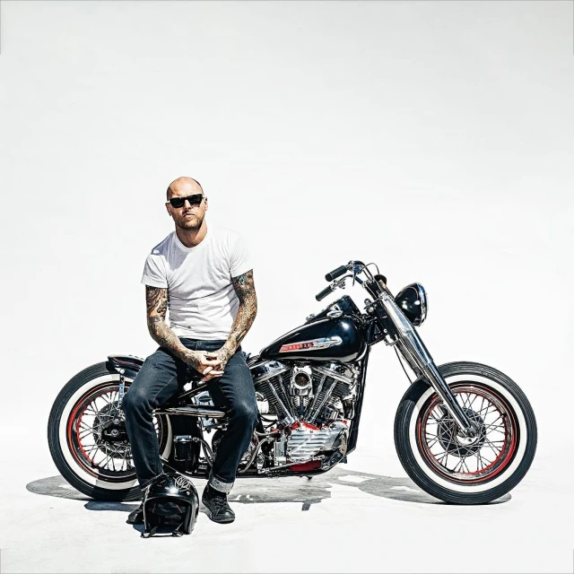 Bald adult male with tattoos sits on a vintage black motorcycle in a bright studio against a white background with a helmet on the ground.