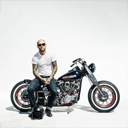 Bald adult male with tattoos sits on a vintage black motorcycle in a bright studio against a white background with a helmet on the ground.