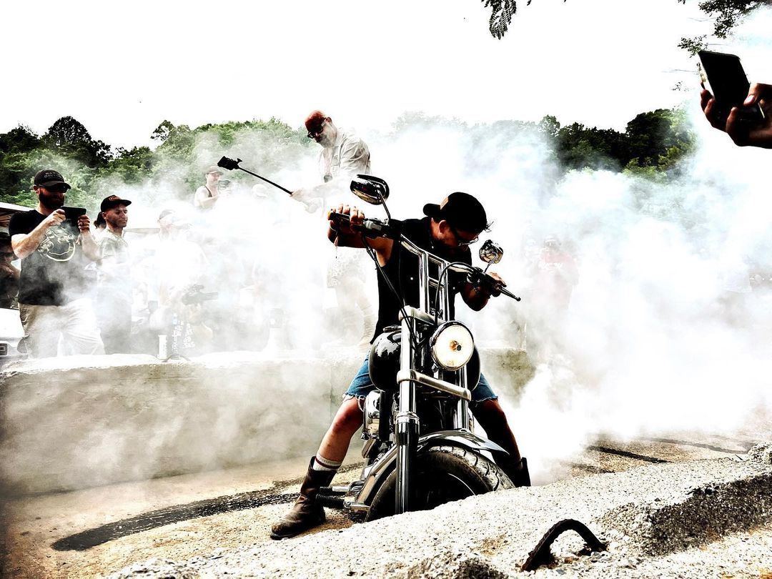 Motorcyclist performing a burnout surrounded by smoke with people filming in an outdoor environment during the daytime.