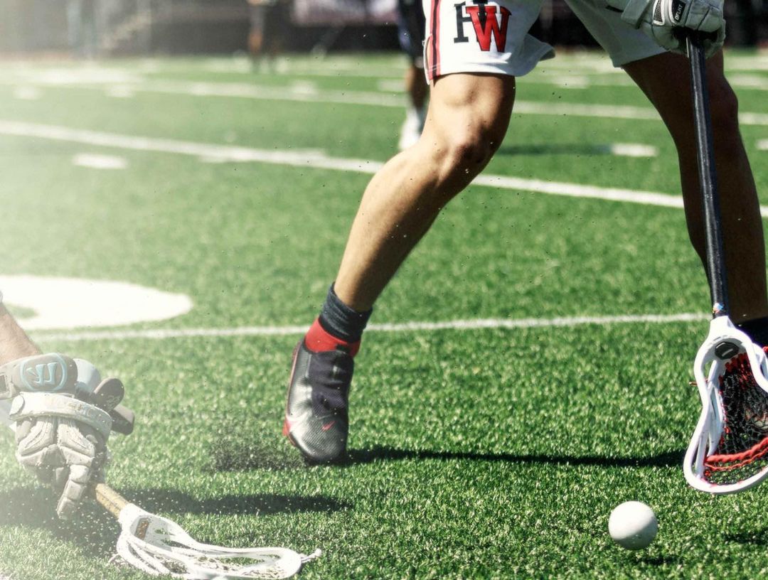 Close-up of a lacrosse player's legs in action on a green field, holding a stick with a ball on the ground during the daytime.