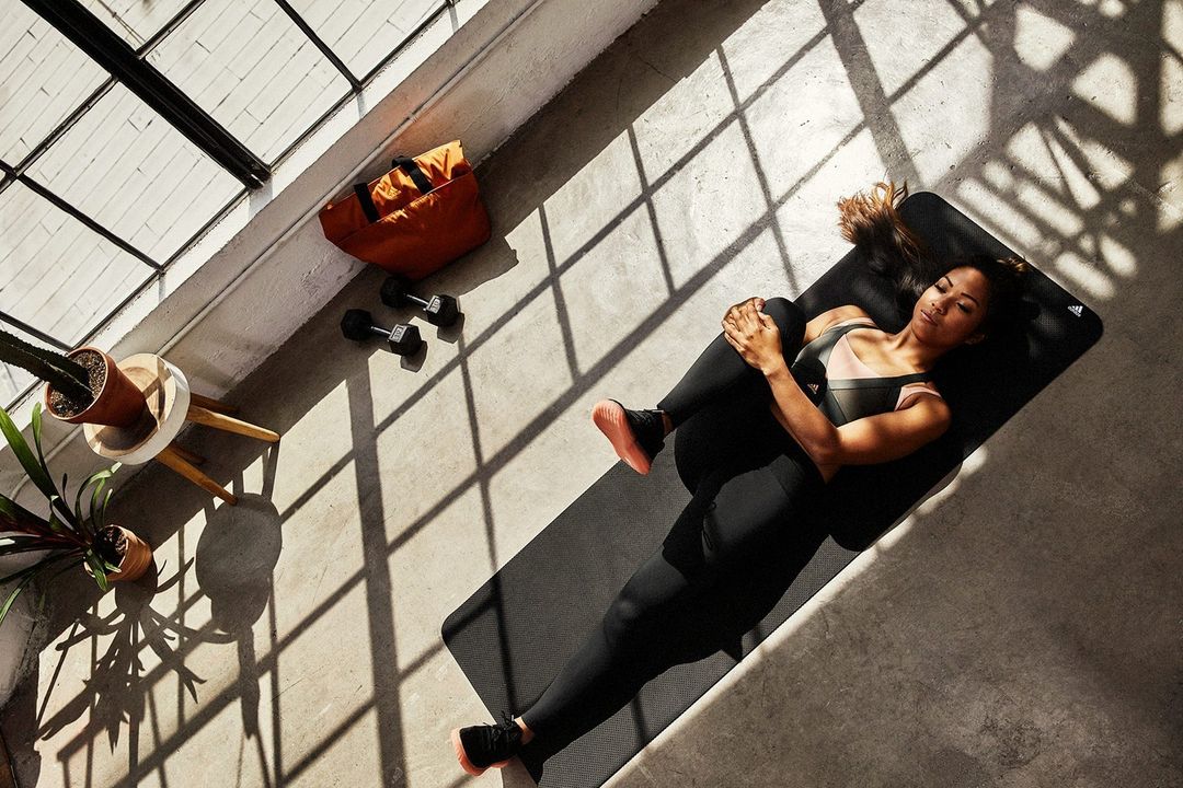 Overhead shot of a woman on a yoga mat in sportswear, stretching with dumbbells, a gym bag, and potted plants nearby in sunlight.