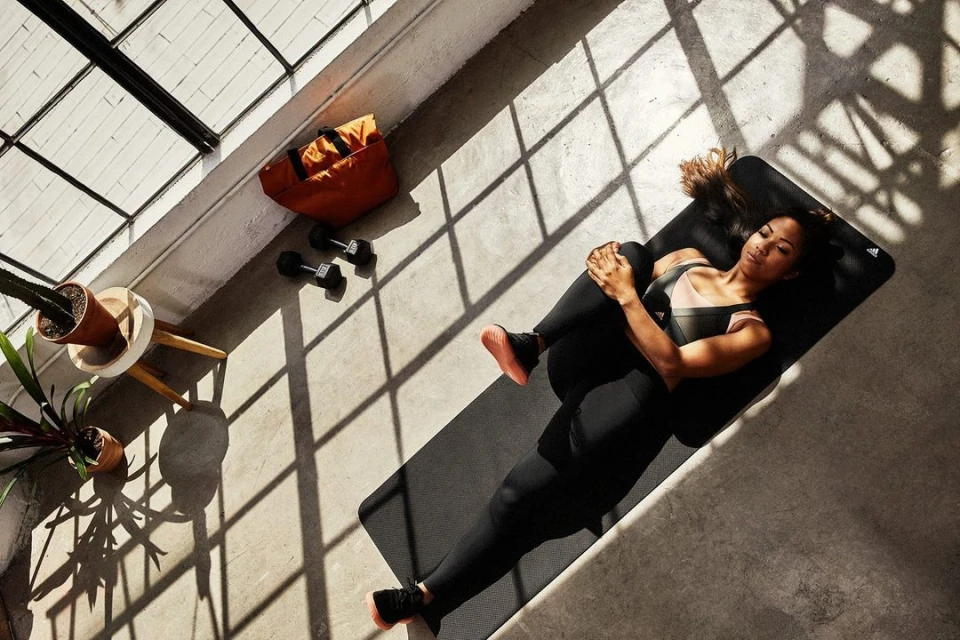 Overhead shot of a woman on a yoga mat in sportswear, stretching with dumbbells, a gym bag, and potted plants nearby in sunlight.