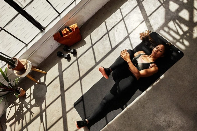 Overhead shot of a woman on a yoga mat in sportswear, stretching with dumbbells, a gym bag, and potted plants nearby in sunlight.