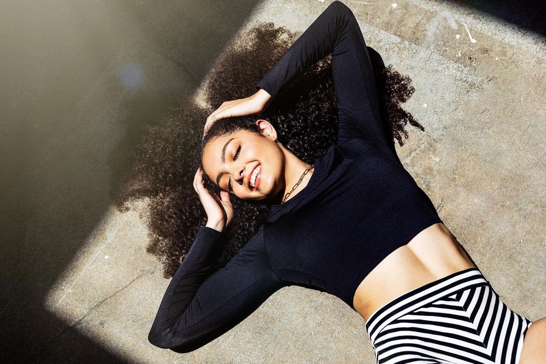 Smiling young woman with curly hair lies on concrete in sunlight wearing a black long-sleeved crop top and patterned shorts.