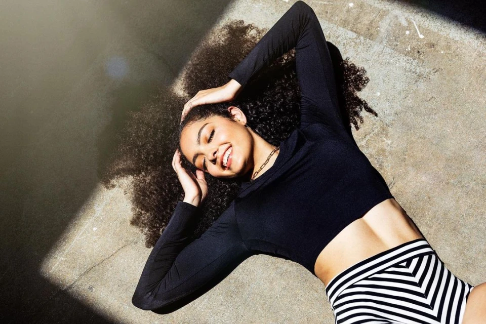 Smiling young woman with curly hair lies on concrete in sunlight wearing a black long-sleeved crop top and patterned shorts.