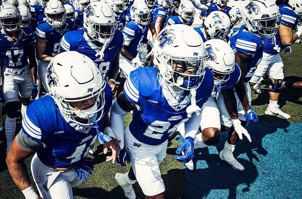 American football players in blue and white uniforms run onto a green field, captured in a medium shot with natural lighting.