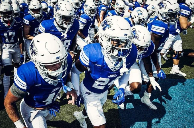 American football players in blue and white uniforms run onto a green field, captured in a medium shot with natural lighting.
