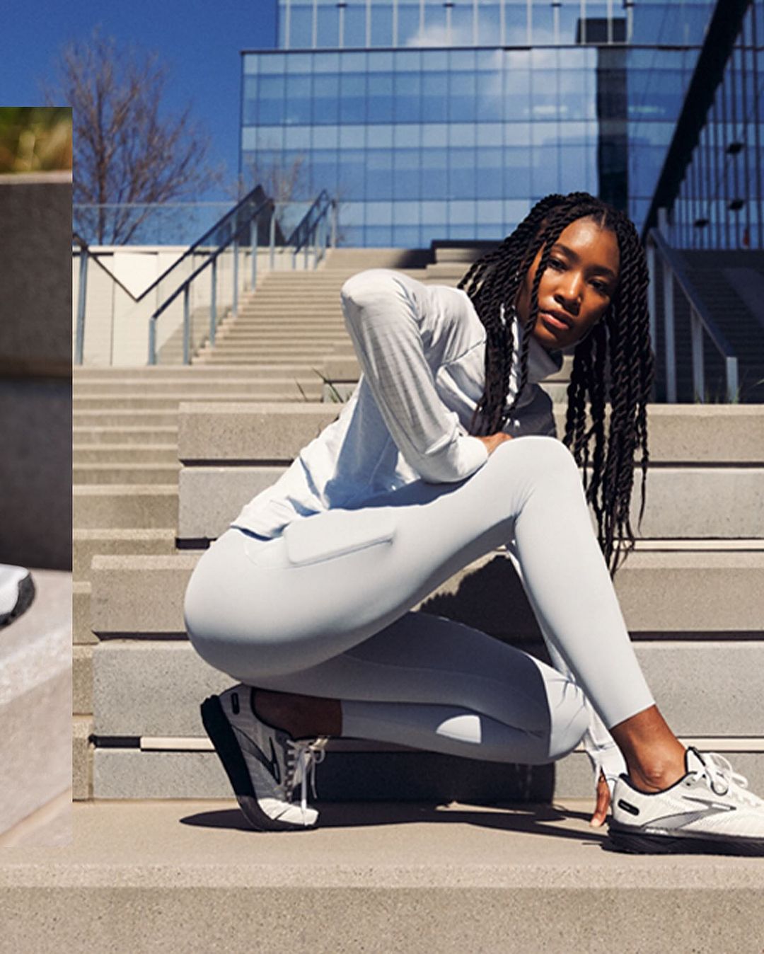 Athletic African-American woman crouches on gray stairs in a light gray long-sleeve top and leggings, wearing white running shoes near a building.