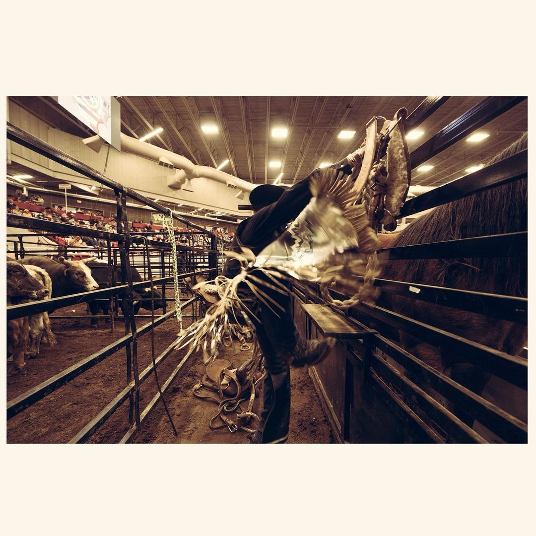 Indoor rodeo event featuring a cowboy preparing equipment in a pen, with blurred audience in the background, under bright arena lighting.