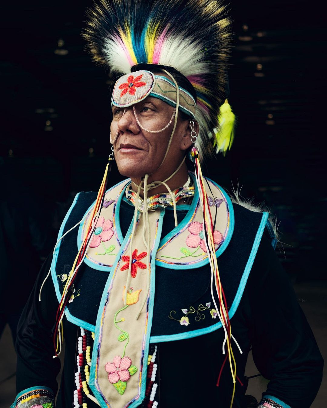 Portrait of a Native American man wearing traditional clothing, including an ornate headdress, beaded adornments, and colorful floral designs.