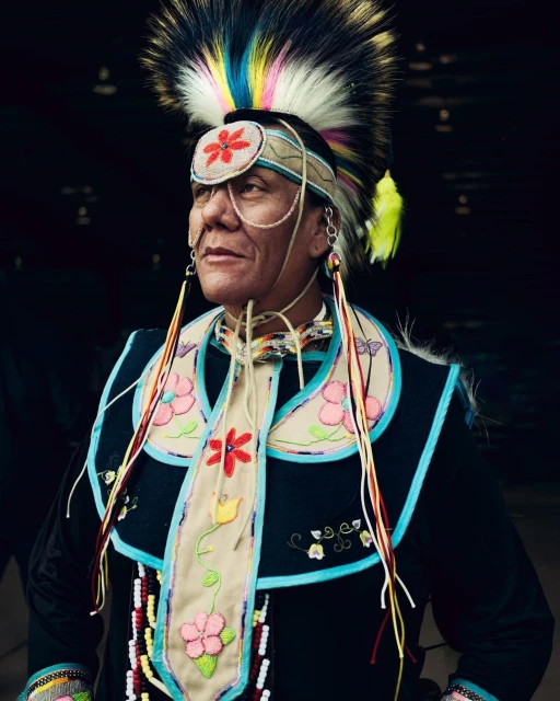 Portrait of a Native American man wearing traditional clothing, including an ornate headdress, beaded adornments, and colorful floral designs.