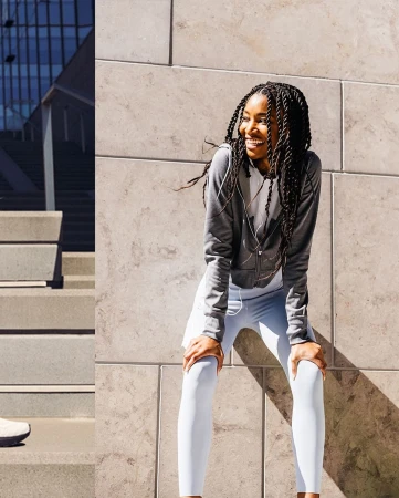A smiling African American woman pauses from her outdoor fitness routine, wearing sporty white leggings and a gray sweatshirt, leaning forward.