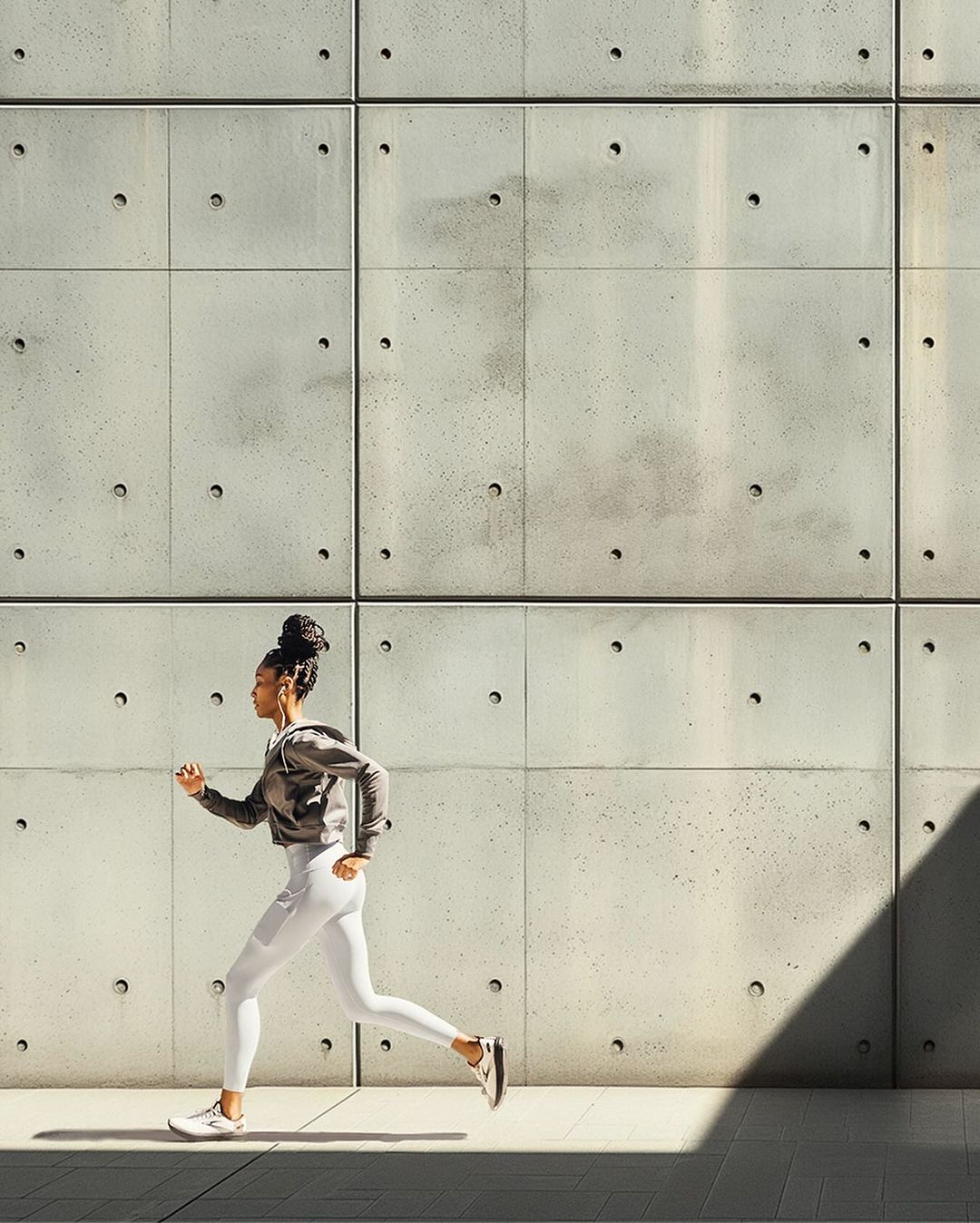 Adult woman with earphones running outdoors against a concrete wall in athletic wear, side view, in natural light.