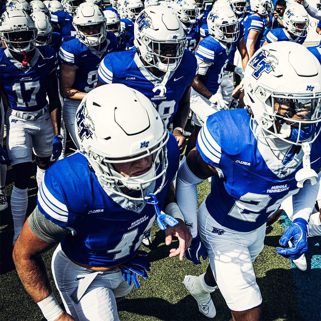 A large group of American football players in blue and white uniforms are running onto the field, viewed from a low angle.
