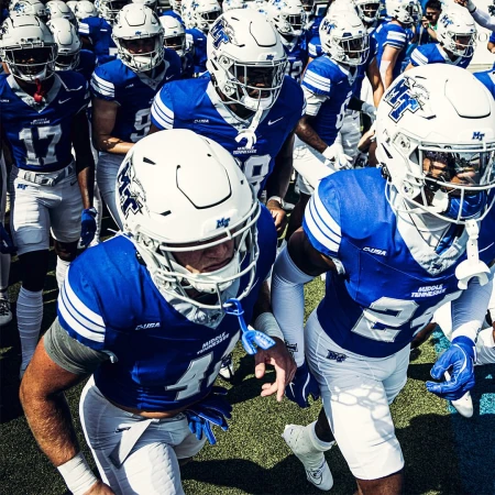 A large group of American football players in blue and white uniforms are running onto the field, viewed from a low angle.